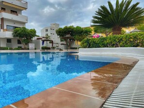 A swimming pool with clear blue water, surrounded by a tiled deck and lush greenery. - Cristian Sur 47 - One Bed (Los Cristianos)