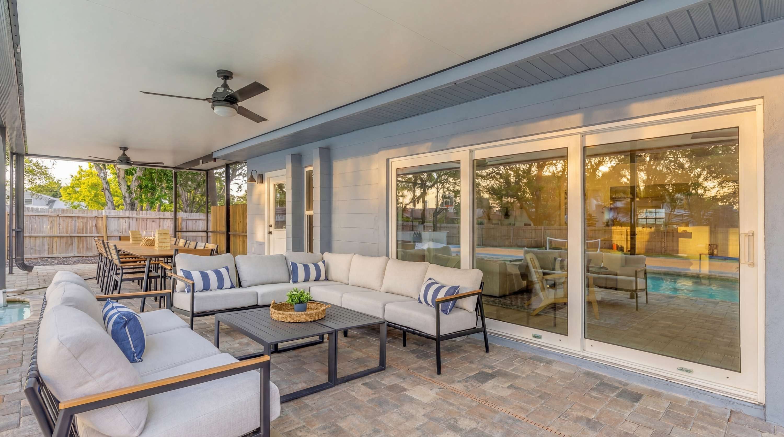 A covered patio area with a sofa set, dining table, and chairs, a ceiling fan, and a view of a swimming pool through glass doors.