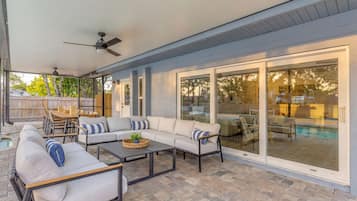 A covered patio area with a sofa set, dining table, and chairs, a ceiling fan, and a view of a swimming pool through glass doors.