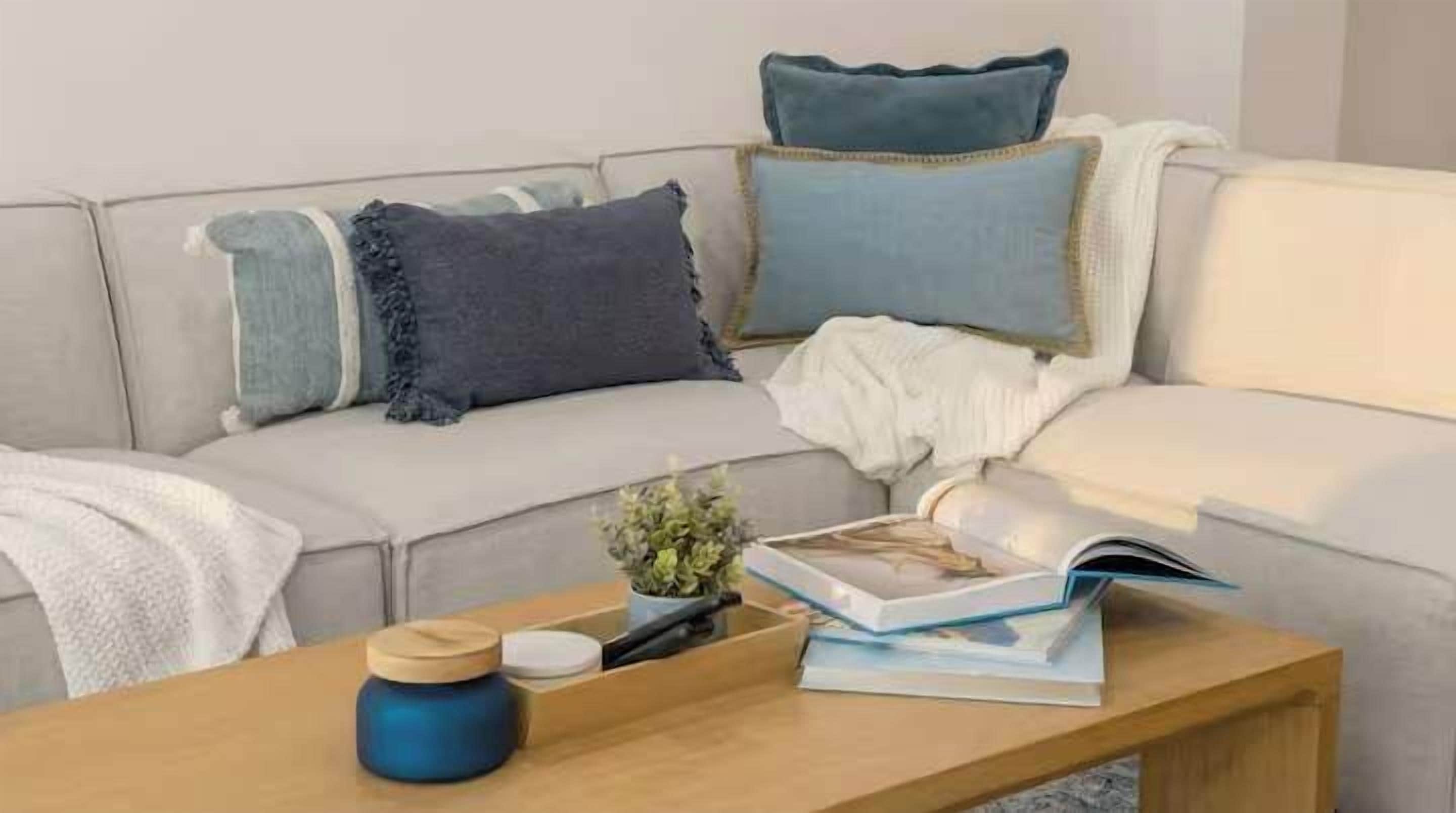 Una sala de estar con un sofá, cojines, una planta pequeña, un libro y una vela sobre una mesa de madera.