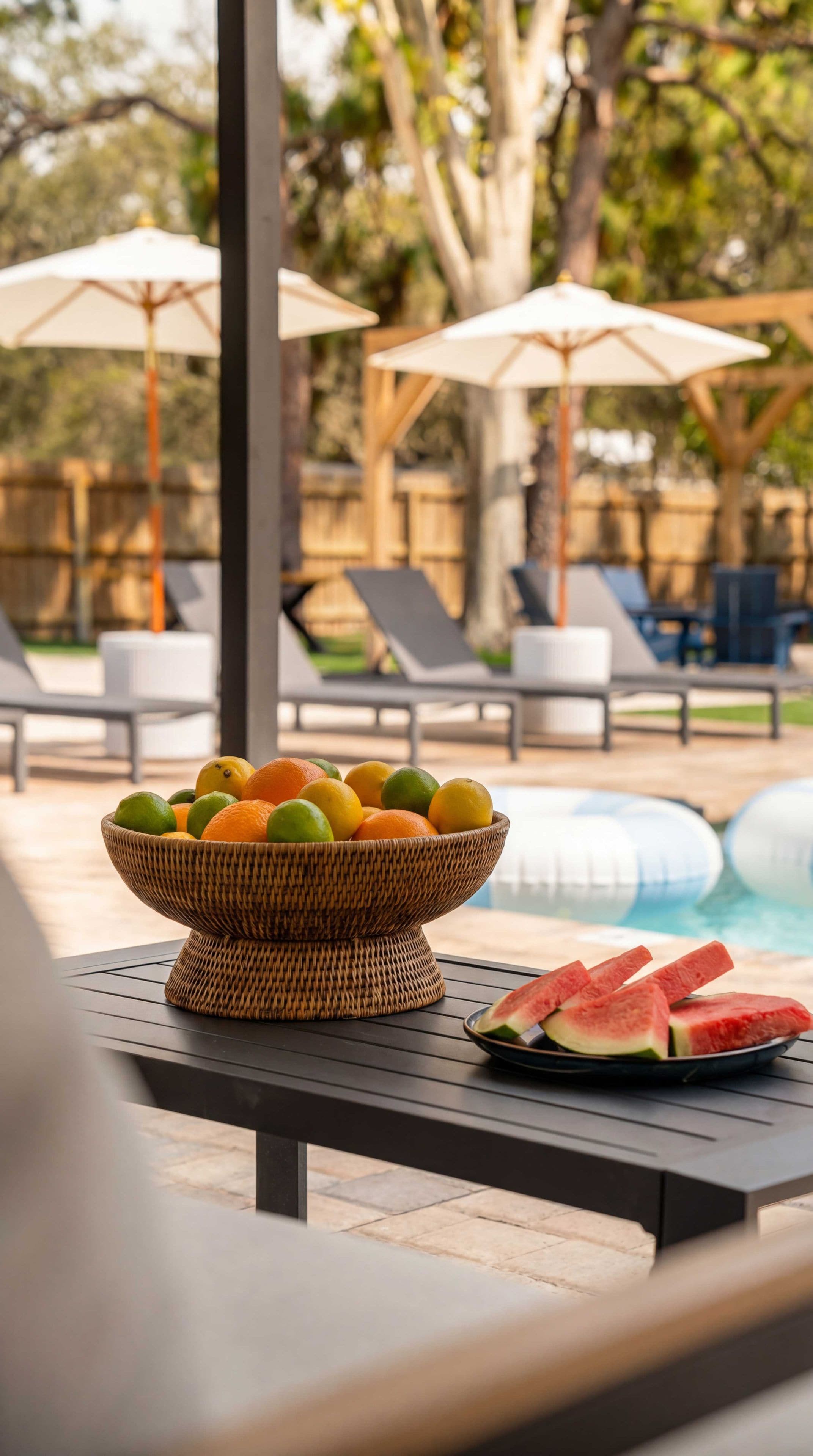 Un tazón de mimbre lleno de fruta sobre una mesa junto a una piscina, con sillones de descanso y sombrillas al fondo.