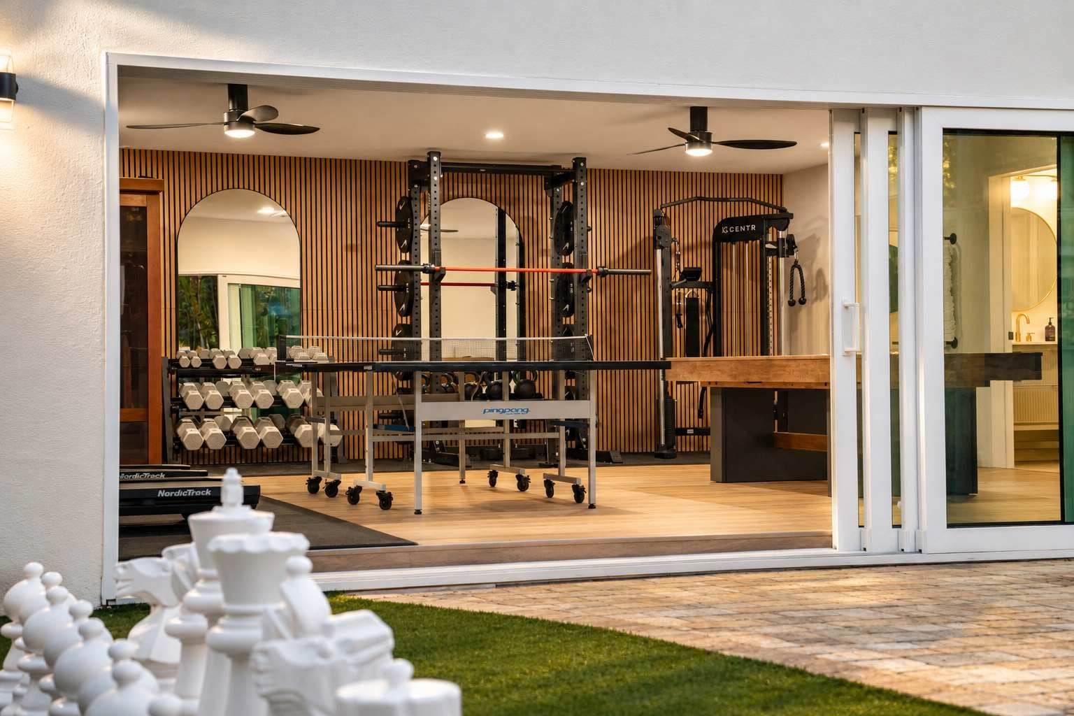 Une salle de sport moderne, équipée de matériel d’exercice, avec un coin repas et une porte-fenêtre coulissante donnant sur un patio.