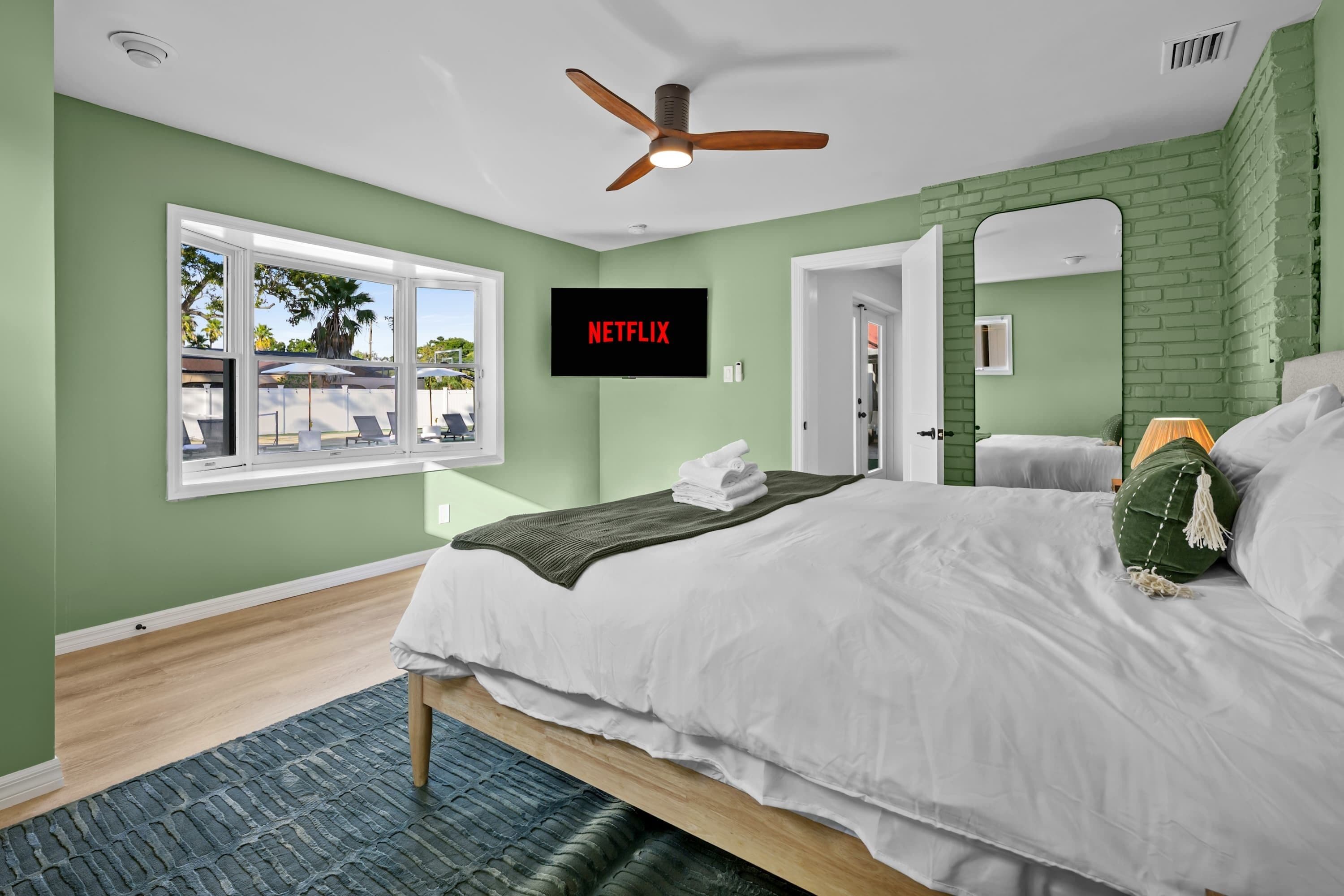 A bedroom with a bed, a ceiling fan, a TV mounted on the wall, and a window with a view of outdoor furniture.