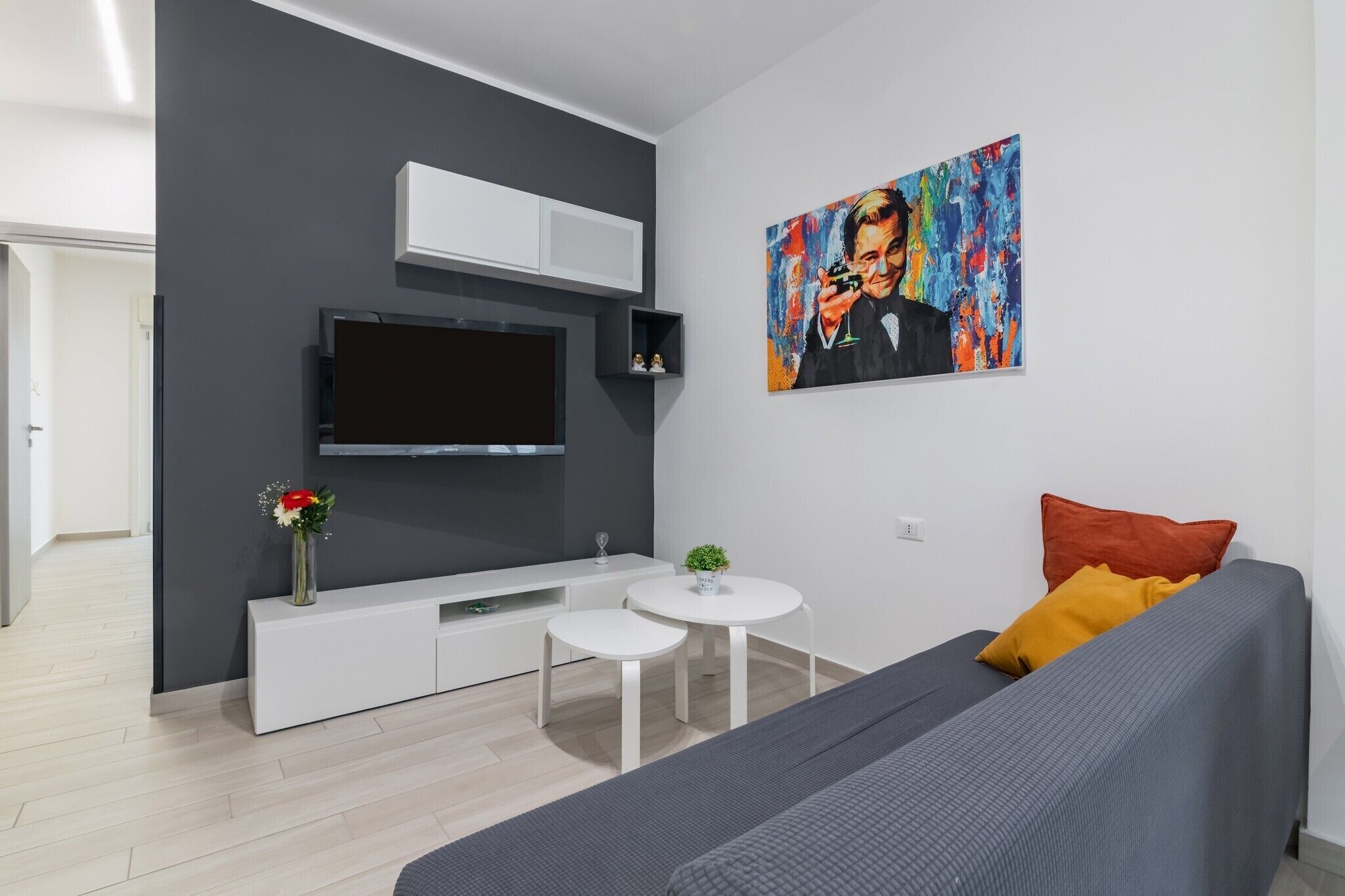 Un salon moderne avec un canapé gris, un téléviseur à écran plat fixé sur un meuble blanc, une petite table ronde ornée d’une plante et un tableau abstrait aux couleurs vives accroché au mur.