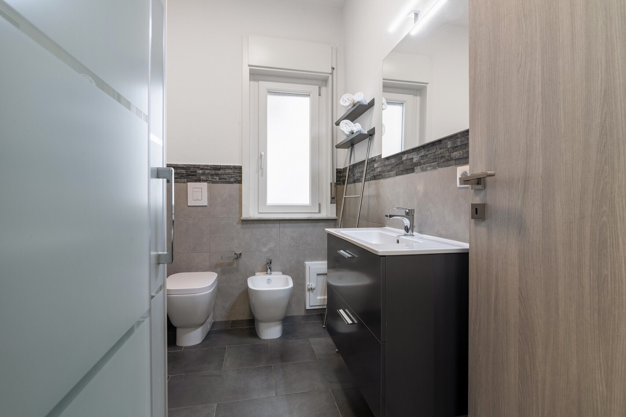 A modern bathroom with a toilet, bidet, vanity, and a window.