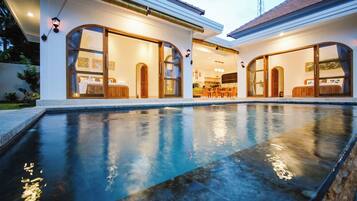 Une maison moderne dotée d’une piscine, de grandes portes vitrées donnant sur un espace repas et d’une cuisine visible à travers le verre.