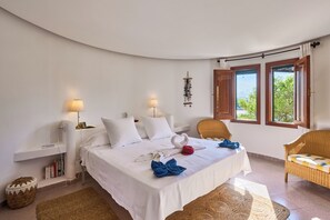 A hotel room with a bed, two bedside lamps, a wicker chair, a small table, and a view of the outdoors through large windows. - Villa Ramina located on the seafront in the old town of Cala Dor. (Santanyí)