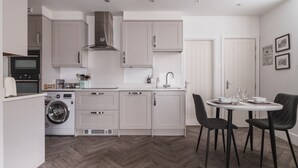 A compact kitchen with a washing machine, a dining area with two chairs, and a herringbone-patterned floor. - Old Village Shop, Ambleside, central, cosy retreat (Ambleside)