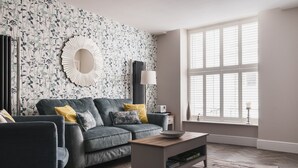 A living room with a grey sofa, a coffee table, and a bookshelf. A large window with blinds is on the right, and a floral wallpaper is on the wall. - Old Village Shop, Ambleside, central, cosy retreat (Ambleside)