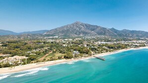Uma cidade costeira com uma montanha imponente ao fundo, uma praia e um píer que se estende para o mar.