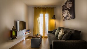 A living room with a grey sofa, a TV mounted on the wall, a bookshelf with books, a decorative lantern, and a large window with yellow curtains. - Lovely cottage for 2 people, roof terrace, 10 minutes from the beach! (Ojén)