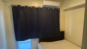 Une petite chambre avec un lit simple, une fenêtre, un luminaire au plafond et un appareil de climatisation.