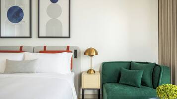 Une chambre d’hôtel avec un lit, un fauteuil vert, une table de chevet avec une lampe et deux tableaux abstraits accrochés au mur.