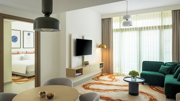 A modern hotel room with a flat-screen TV, a sofa, a dining table with chairs, and a decorative rug. - Bloom Arjaan By Rotana (Abu Dhabi)