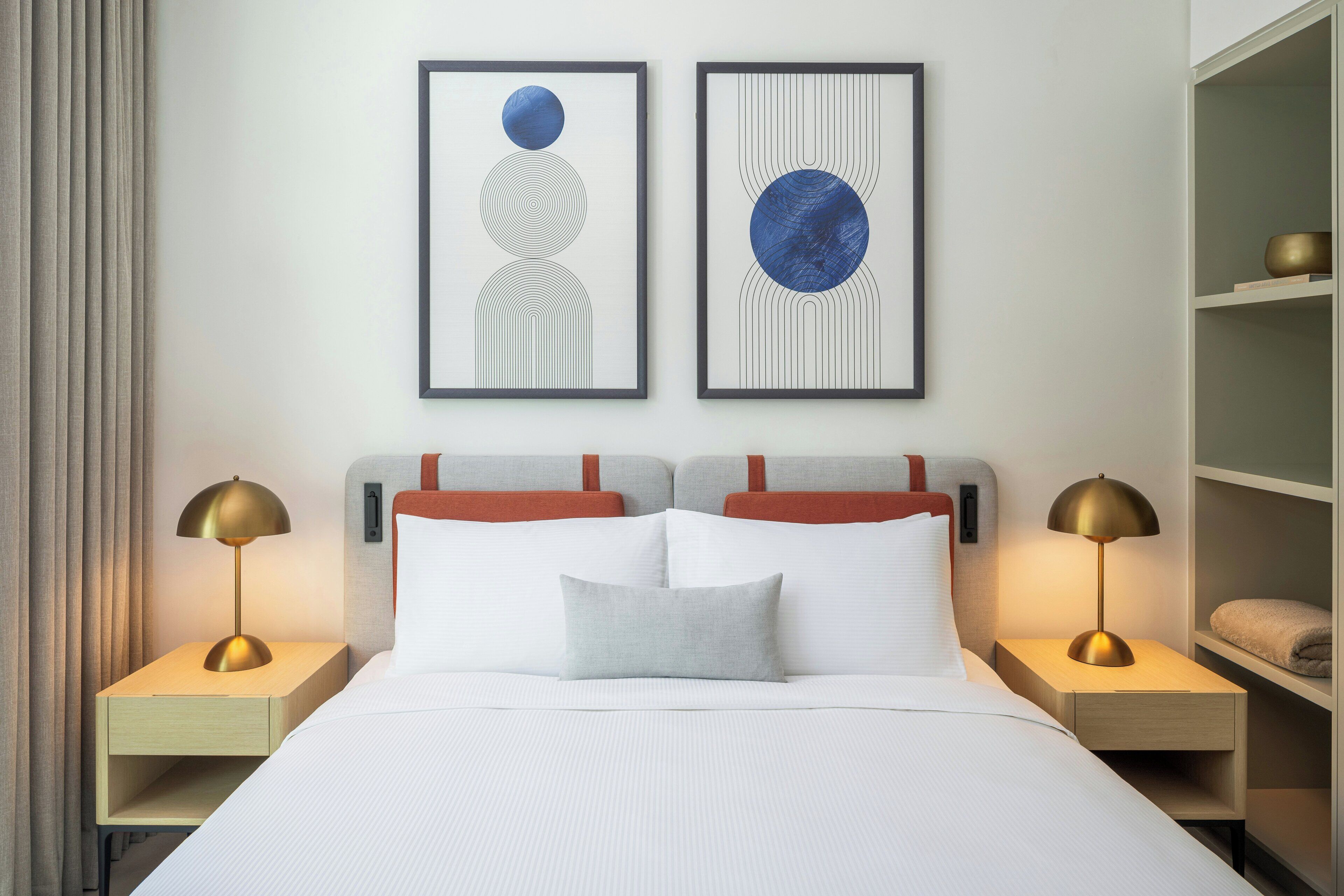 A hotel room with a bed, two bedside tables with lamps, a headboard, and two framed artworks on the wall.