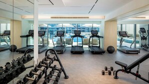 A gym with treadmills, dumbbells, and exercise balls, with large windows and a view of buildings. - Bloom Arjaan By Rotana (Abu Dhabi)
