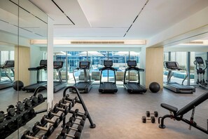 Een fitnessruimte met loopbanden, gewichten en fitnessballen, met grote ramen en uitzicht op gebouwen.