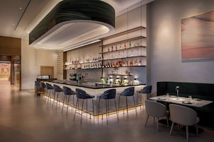 Un área de bar moderna con una barra alargada, varios taburetes y un rincón con mesa y sillas.