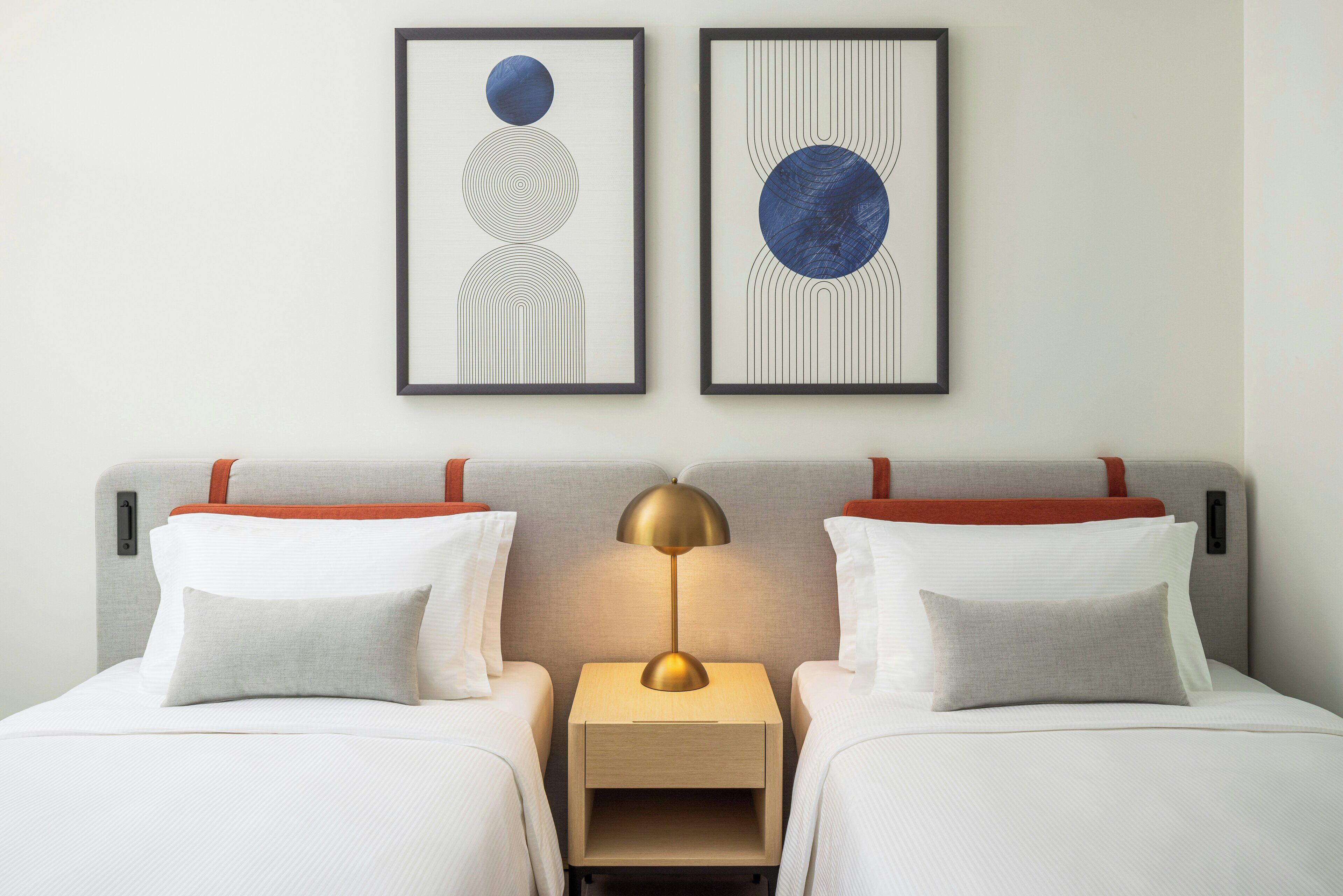 A hotel room with two beds, a nightstand, a lamp, and two framed artworks on the wall.