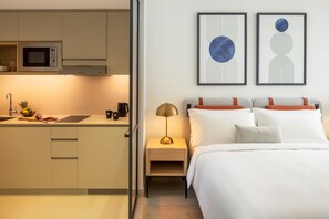 A modern hotel room with a kitchenette, a bed with white bedding, and two abstract art pieces on the wall.