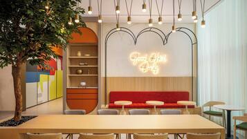A modern restaurant interior with a long wooden table, cushioned chairs, a neon sign, and a decorative tree.