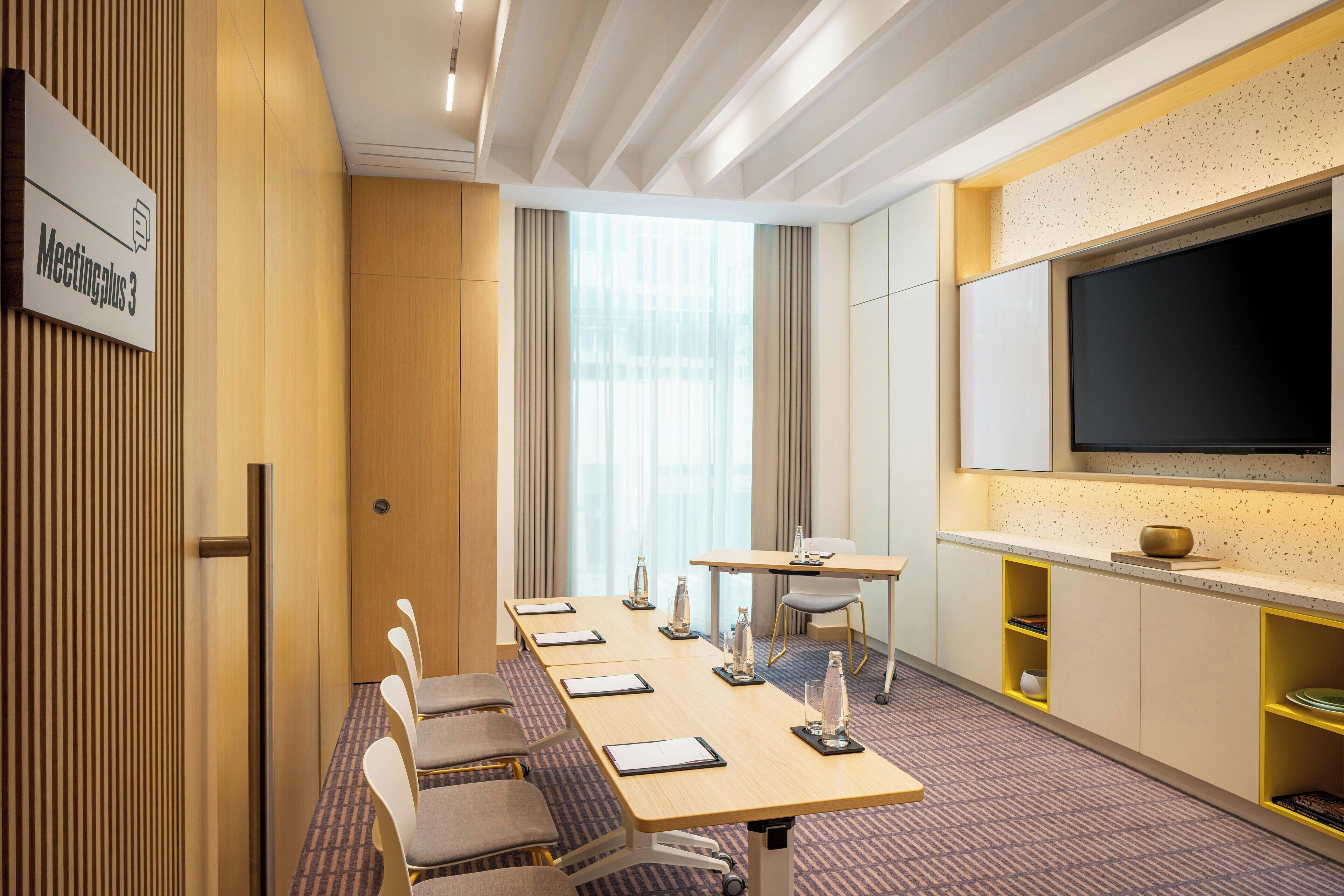 A hotel meeting room with a long table, chairs, a large screen, and a sign indicating the room number.