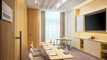 A hotel meeting room with a long table, chairs, a large screen, and a sign indicating the room number.