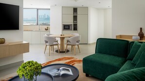 A modern living room with a green sofa, a round dining table with chairs, a flat-screen TV mounted on the wall, and a view of the cityscape through large windows. - Bloom Arjaan By Rotana (Abu Dhabi)