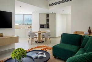 Una sala de estar moderna con un sofá verde, una mesa de comedor redonda con sillas, un televisor de pantalla plana montado en la pared y vistas de la ciudad a través de amplios ventanales.