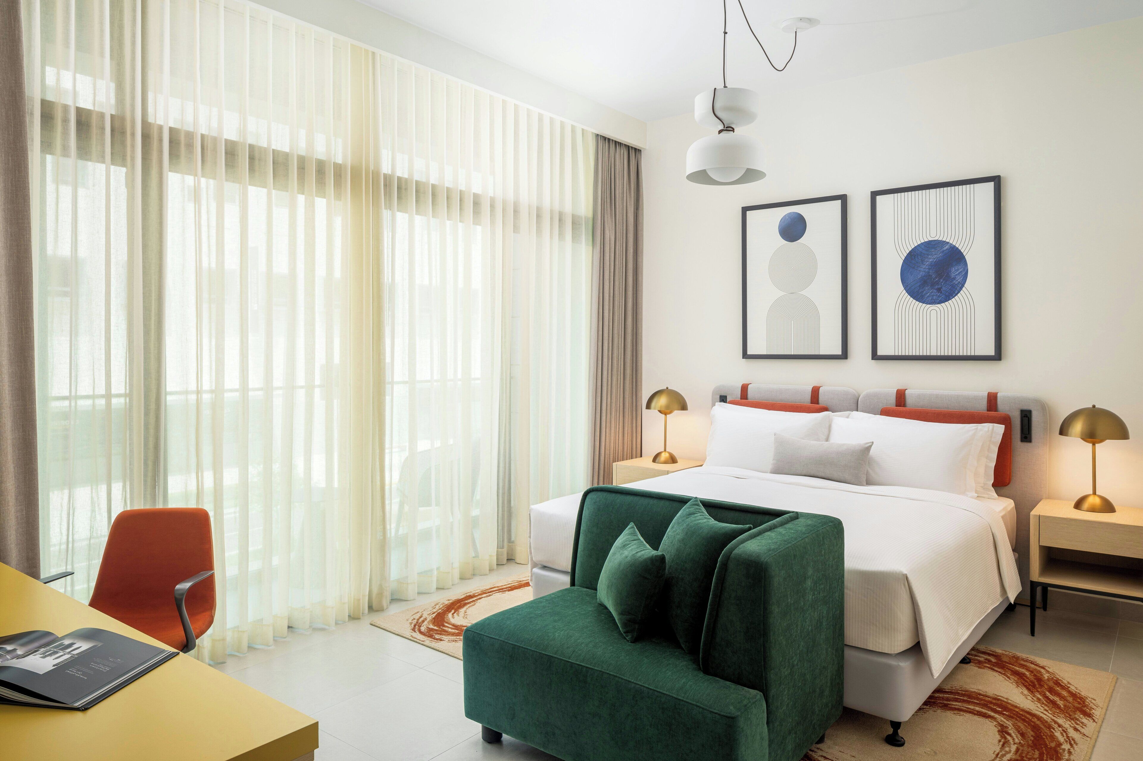 Habitación de hotel con una cama, dos mesitas de noche, un sillón verde, un escritorio con una silla y un ventanal con cortinas traslúcidas.