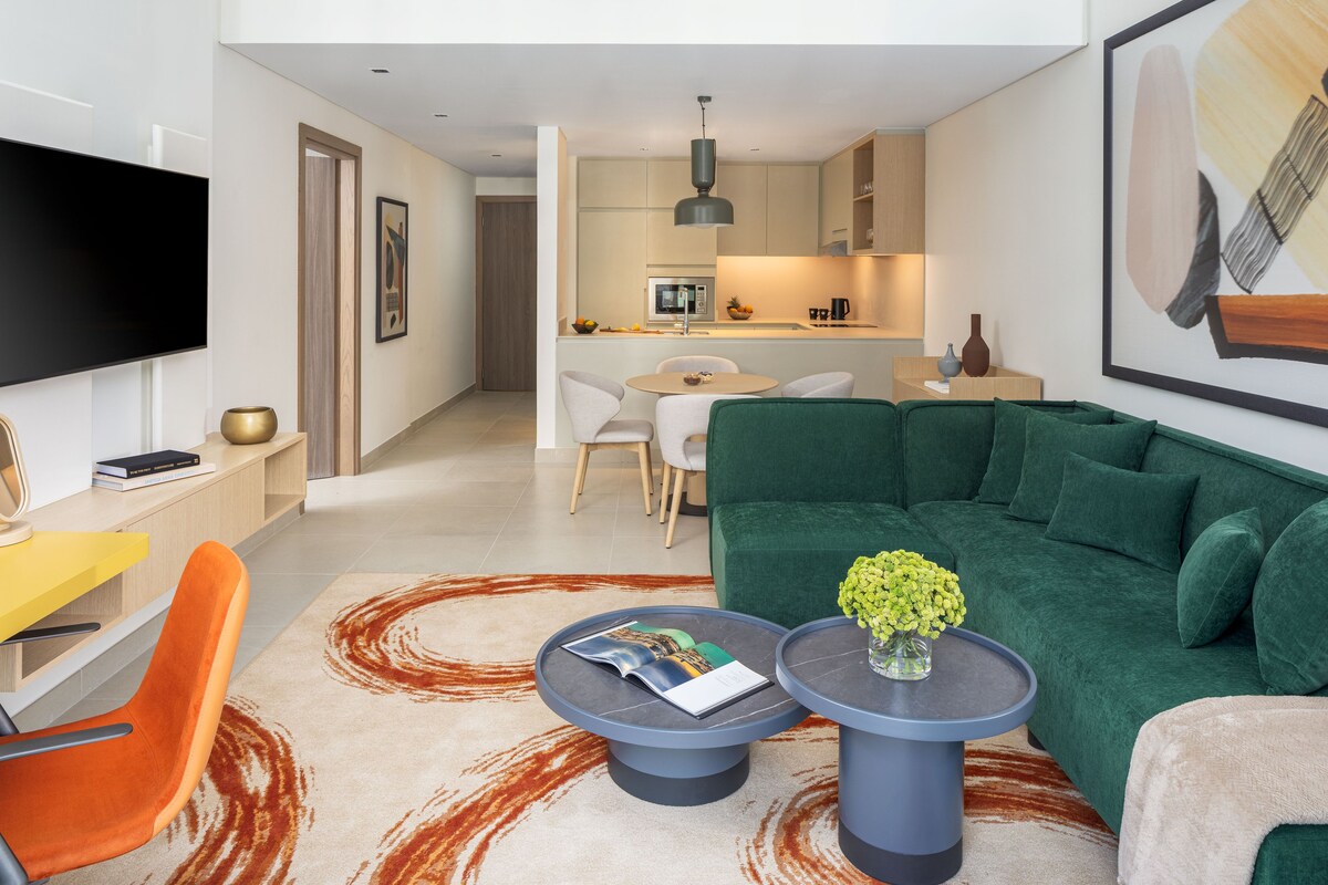Una sala de estar moderna con un sofá verde, una mesa de centro y un televisor. Se ve una cocina al fondo con un comedor.
