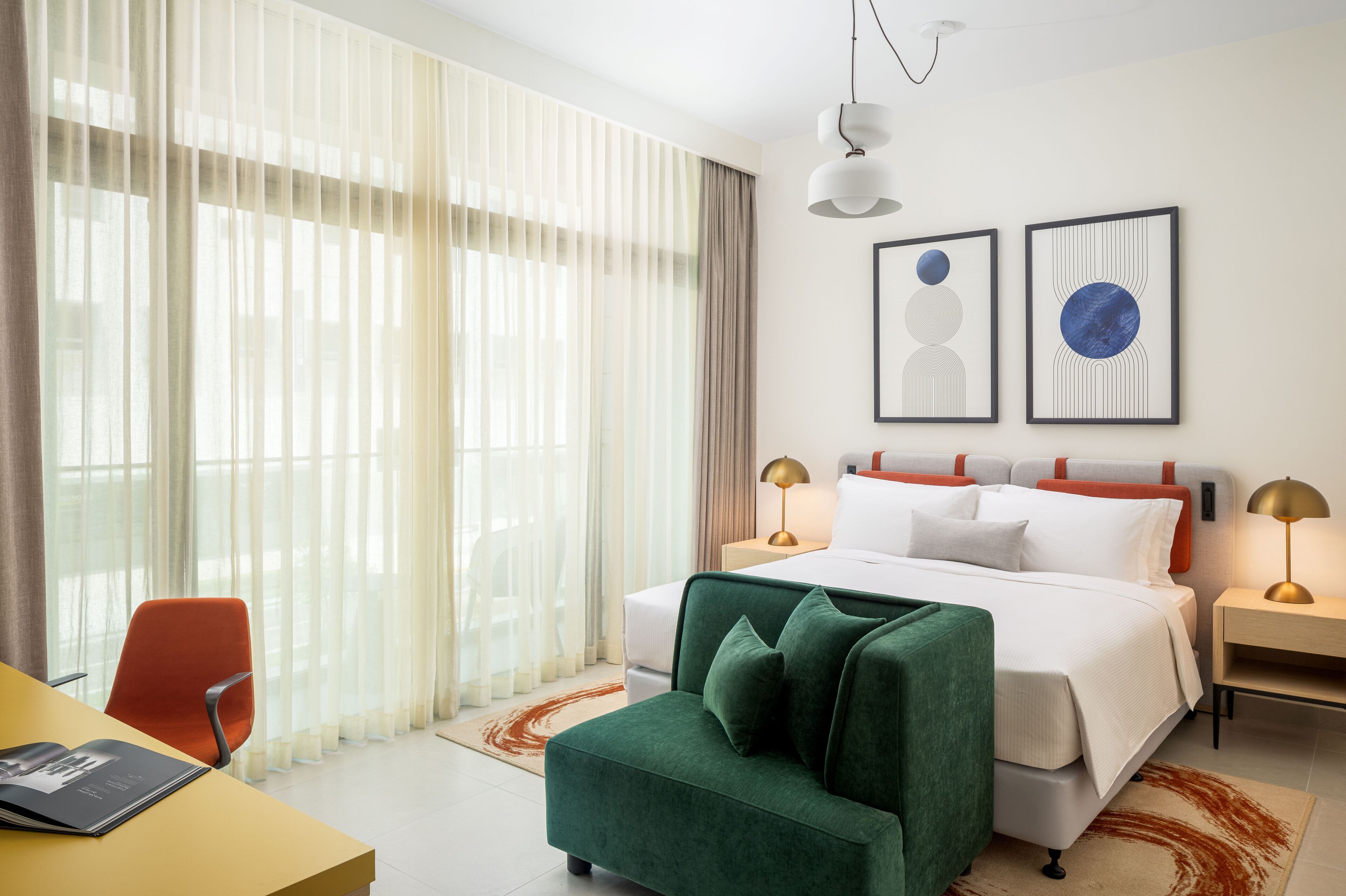 A hotel room with a bed, a green armchair, a desk, and two bedside tables with lamps.