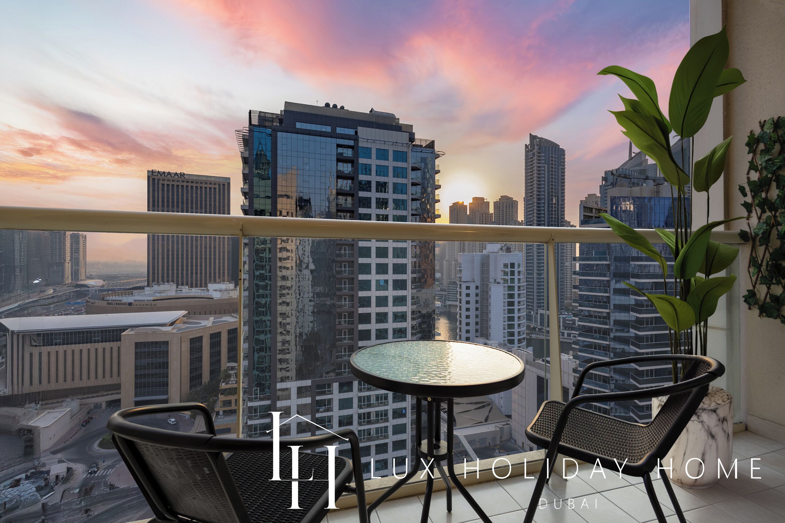 Ein Balkon mit Glastisch und zwei Stühlen mit Blick auf eine Stadtlandschaft, moderne Gebäude und einen Sonnenuntergang.