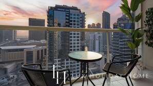 Een balkon met een glazen tafel en twee stoelen, met uitzicht op een stadsgezicht met moderne gebouwen en een zonsondergang.
