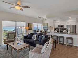 Ein modernes Wohnzimmer mit einem dunkelgrauen Sofa, einem Holztisch und einem gemusterten Sessel. Ein Deckenventilator und Blick aufs Meer sind zu sehen.