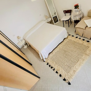 Ein Schlafzimmer mit einem Bett, einem kleinen runden Tisch, einem Stuhl, einem Spiegel und einer Zimmerpflanze.