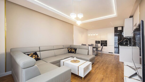 Ein modernes Wohnzimmer mit einem grauen Ecksofa, einem weißen Couchtisch und einer Küche im Hintergrund.