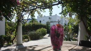 A garden with a white building in the background, a clear sky, and a pathway leading to a glass vase with pink flowers. - Granma Butik Apart Otel (Bodrum)