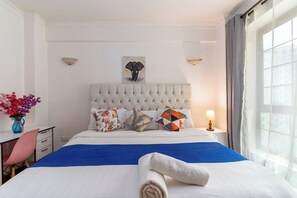 Hotelkamer met een gestoffeerd hoofdeinde, een bed met een blauwe spreideken, meerdere kussens, een nachtkastje met een lamp, een bureau met stoel, en een raam met gordijnen.