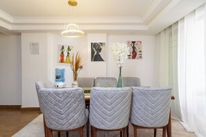 A dining area with a table, three grey upholstered chairs, a water dispenser, and decorative items on the wall. - Luxurious 4 Bedroom Apartment With a Rooftop Pool in Kilimani (Nairobi)