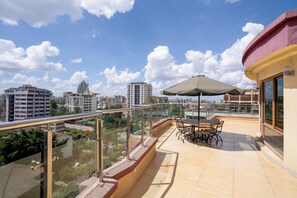 Een balkon met een tafel, stoelen, een glazen balustrade en uitzicht op stadsgebouwen.