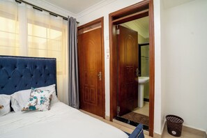 A bedroom with a tufted headboard, a bed with white bedding and floral pillows, a wooden door, and a bathroom visible through an open door. - Luxurious 4 Bedroom Apartment With a Rooftop Pool in Kilimani (Nairobi)