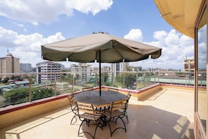 Een balkon met een tafel, stoelen en een grote parasol, met uitzicht op de stad.