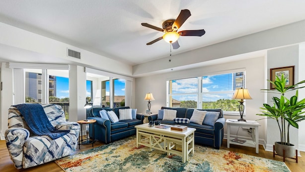 A spacious living room with a ceiling fan, a blue sofa set, a coffee table, and a large window with blinds. - Oceanfront 3BR Condo • Pool & Panoramic Views (New Smyrna Beach)