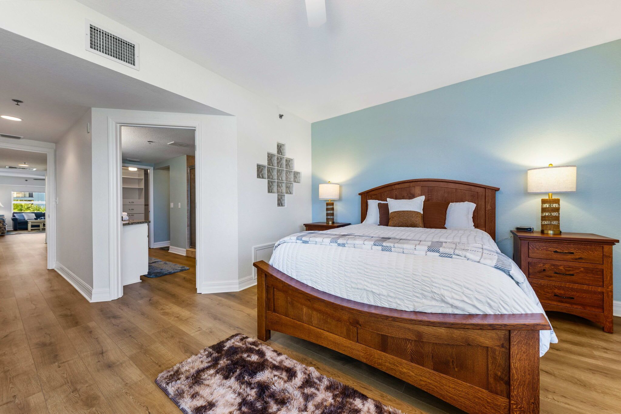 Une chambre à coucher avec un lit en bois, deux tables de chevet avec des lampes, une décoration murale en forme de croix et un couloir menant à une autre pièce.