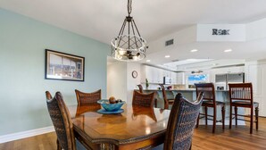A dining area with a wooden table and chairs, a chandelier, and a kitchen in the background. - Oceanfront 3BR Condo • Pool & Panoramic Views (New Smyrna Beach)
