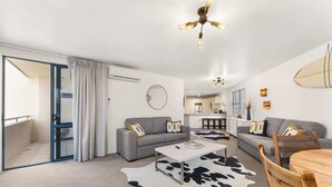 Ein modernes Wohnzimmer mit grauem Sofa, einem weißen Couchtisch und einem Kuhfellteppich. Es gibt einen Essbereich mit Holztisch und Stühlen. Der Raum verfügt über einen Deckenventilator und eine Leuchte.
