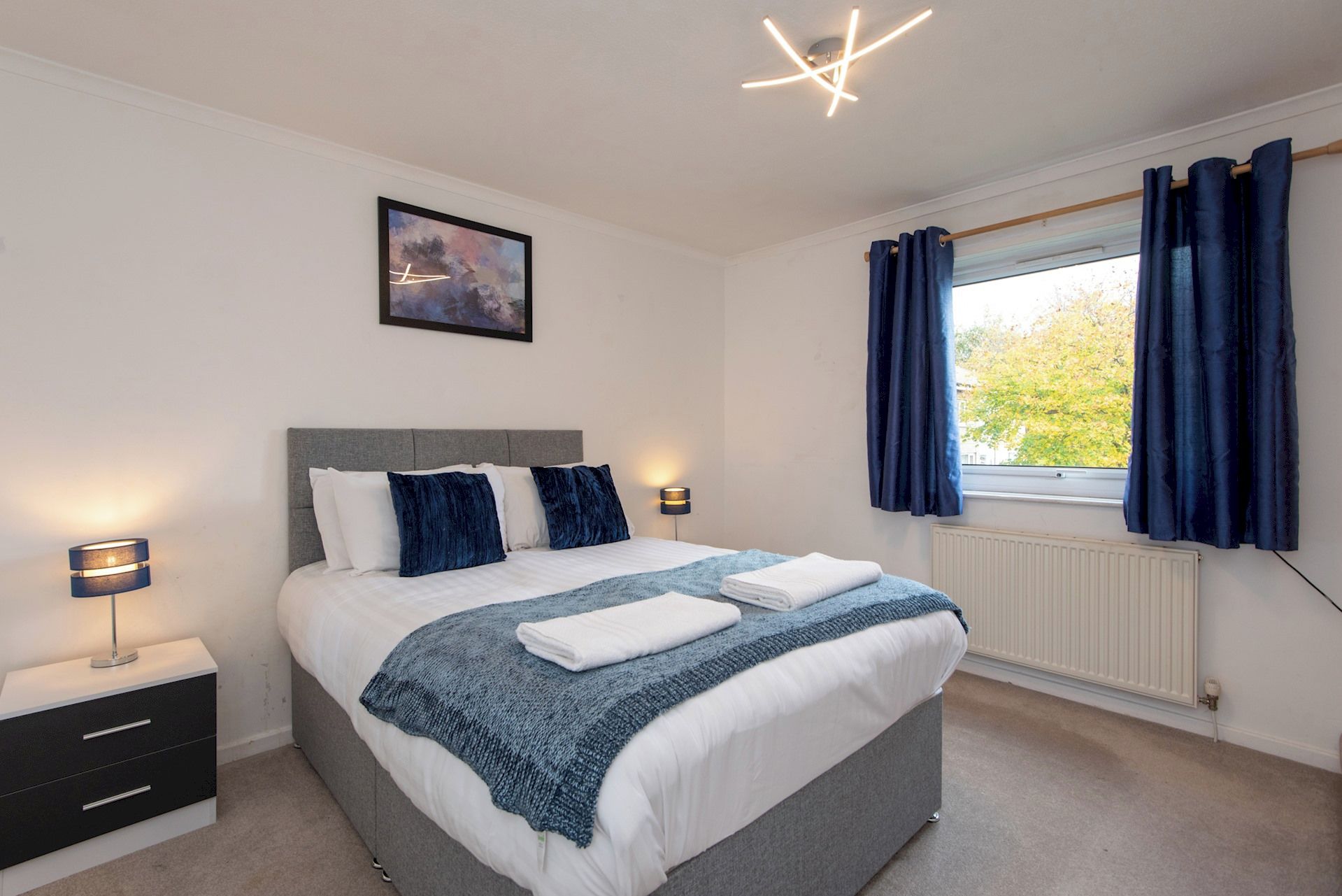 A modern bedroom with a bed, bedside tables, a window with curtains, and a wall-mounted light fixture.