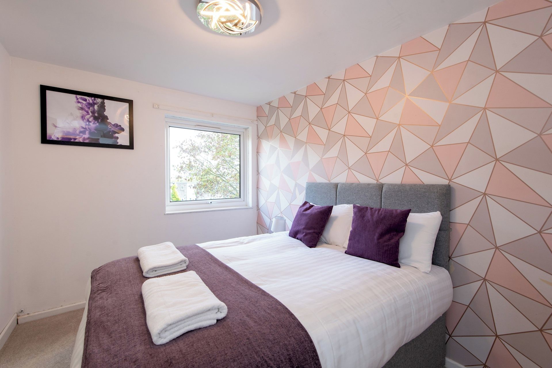 A bedroom with a bed, pillows, a blanket, a window, and a geometric patterned wall.