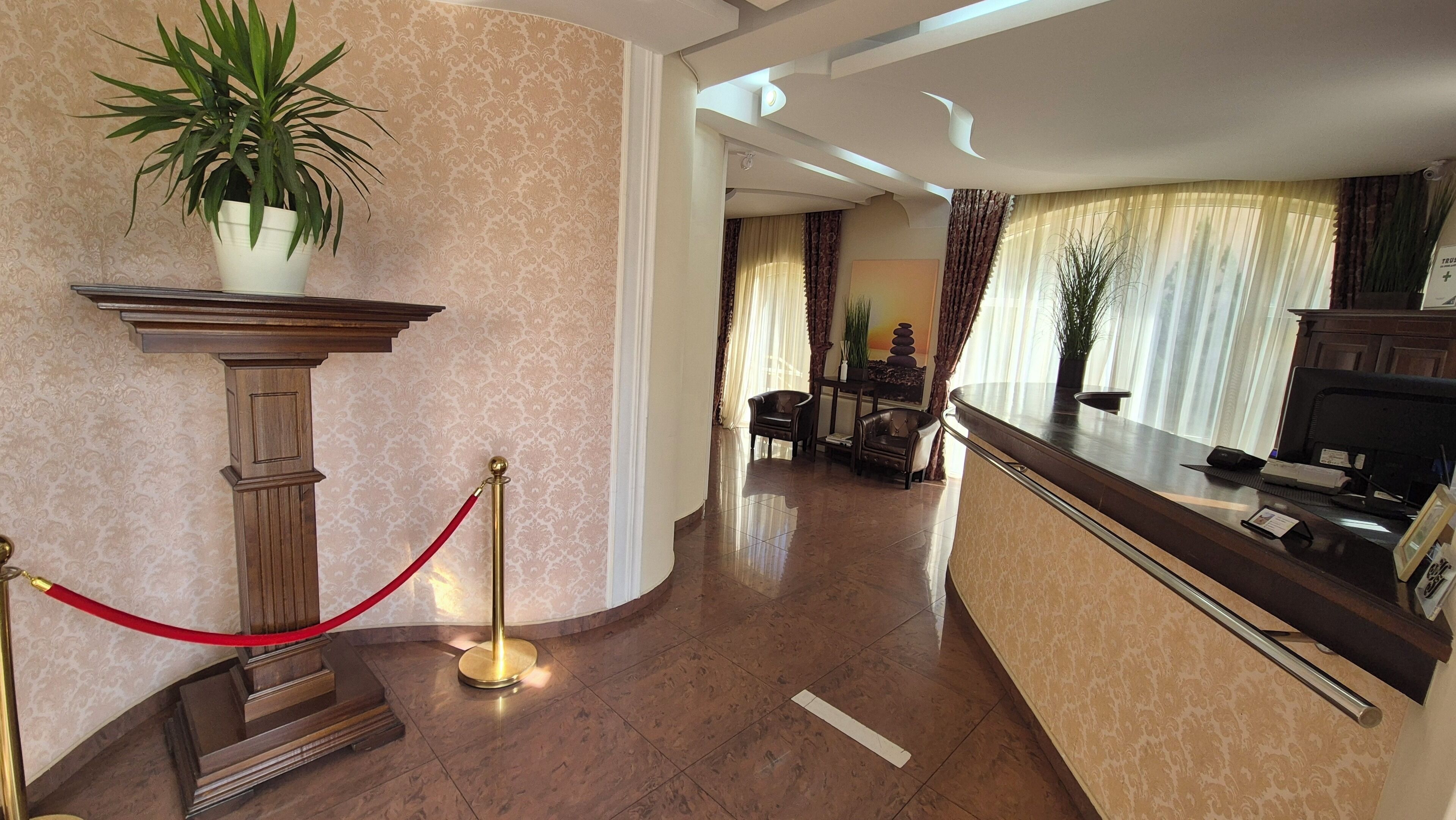Un hall de hotel con mostrador de recepción, una barrera de cuerda roja, una planta en maceta y una escalera que lleva a otra zona.